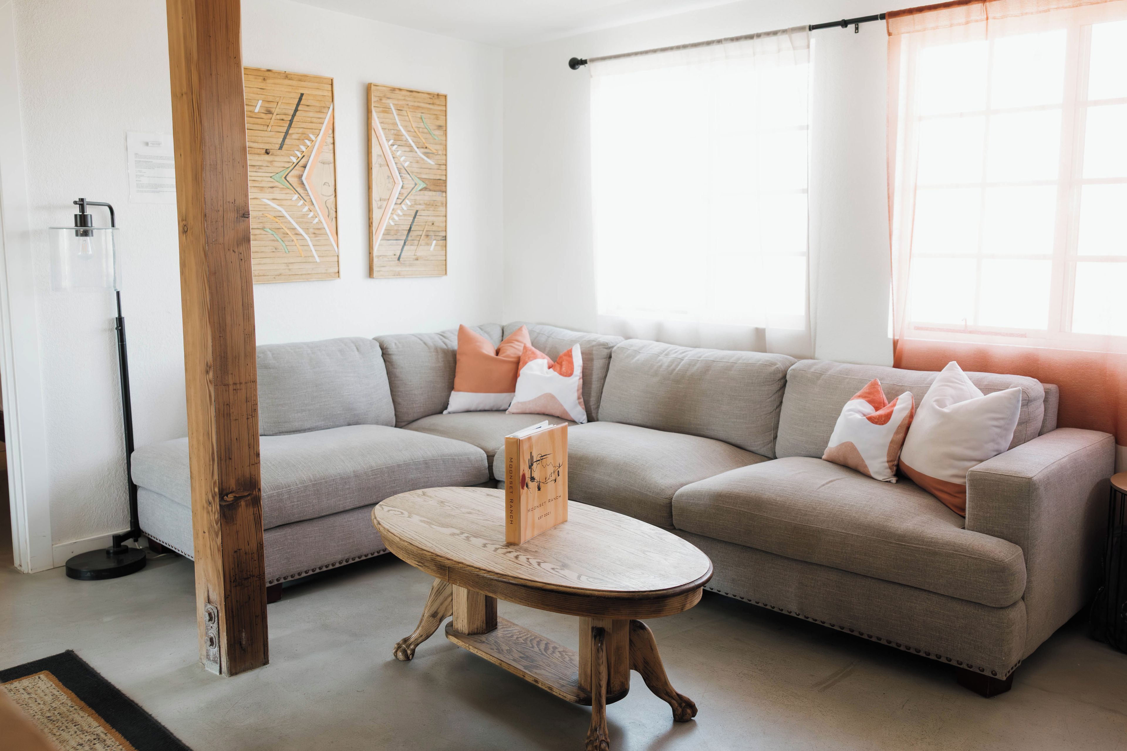 Un salon moderne avec un canapé d’angle gris, une table basse ronde en bois et une petite table d’appoint. Deux œuvres d’art sont accrochées au mur, et une fenêtre est agrémentée de rideaux légers.