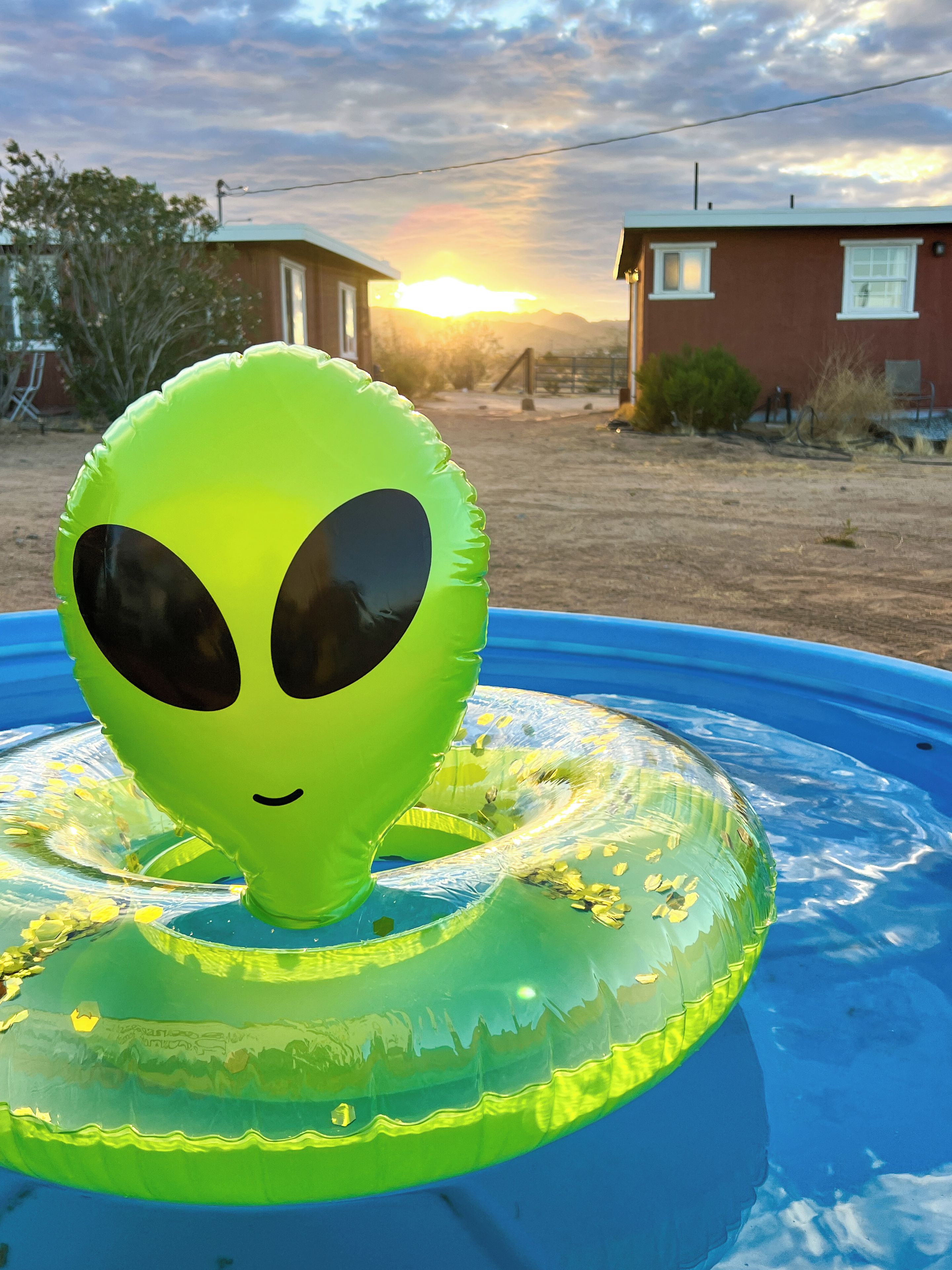 Una cara de extraterrestre inflable sobre una piscina inflable azul, con un atardecer de fondo.