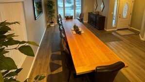 Un comedor amplio con una larga mesa de madera, varias sillas, una planta en maceta y vistas a través de grandes ventanales.