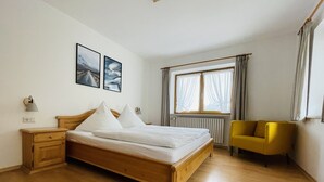 Een slaapkamer met een houten bed, twee nachtkastjes, een raam met gordijnen en een gele fauteuil.