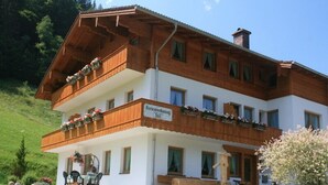 A two-story wooden building with a balcony, a sign reading "Ferienwohnung Drei", and a log sculpture in front.