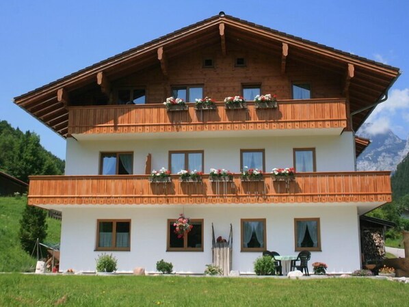 Un chalet en bois à deux étages, doté de balcons et d’une façade blanche, avec une vue sur les montagnes en arrière-plan.