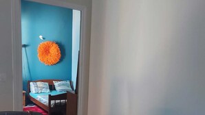 A hallway with a blue wall, a wooden door, and a bed with a red blanket in the background. - Studio nono (Pointe-à-Pitre)