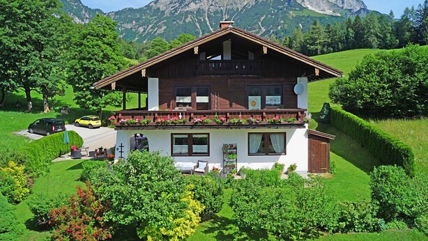 Een chalet met balkon, omringd door weelderig groen en bergen op de achtergrond.