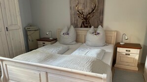 A bedroom with a bed, two bedside tables, a nightstand, a lamp, a wall-mounted artwork of a deer, and a door.