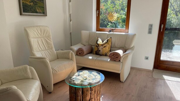 A living room with a beige sofa, a matching armchair, and a glass coffee table on a wooden stump. A window with a sun decoration is visible.