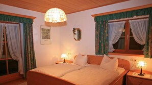 A cozy bedroom with a wooden bed, two bedside tables with lamps, a hanging light fixture, and curtains on the windows. - Apartment 4 for 2 persons with balcony, 27 sqm, kitchen, living room/bedroom (Inzell)