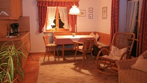 A cozy dining area with a table, chairs, and a bench. There is a window with curtains, a lamp, and a plant. - Apartment 4 for 2 persons with balcony, 27 sqm, kitchen, living room/bedroom (Inzell)