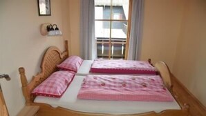Ein hölzernes Bett mit zwei rosa karierten Bettlaken und Kissen, ein hölzernes Kopfteil und ein Fenster mit Vorhängen.