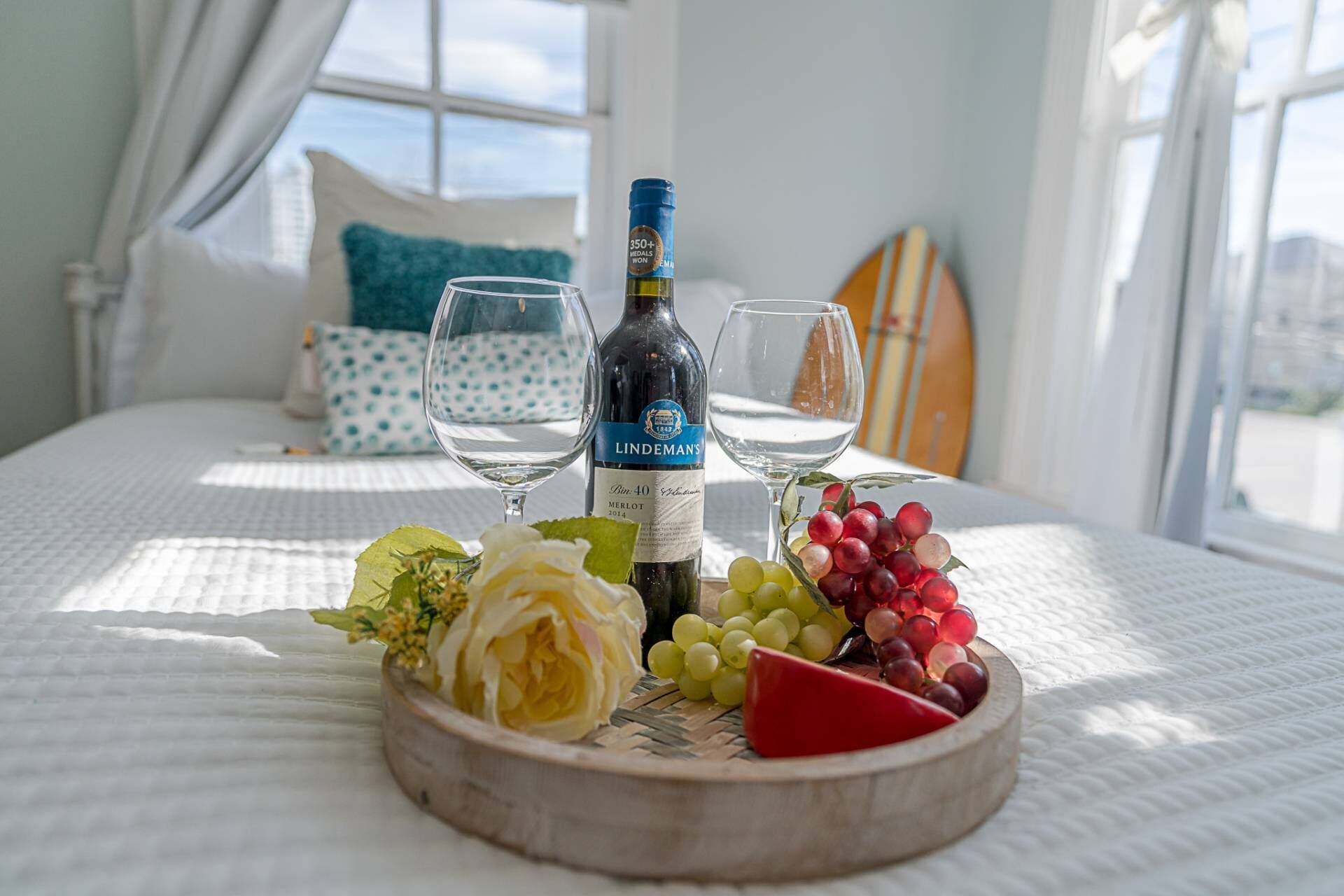 A bed with a white bedspread, a bottle of wine, two glasses, and a wooden board with cheese, grapes, and a rose.