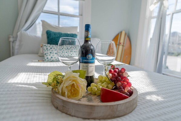 A bed with a white bedspread, a bottle of wine, two glasses, and a wooden board with cheese, grapes, and a rose.