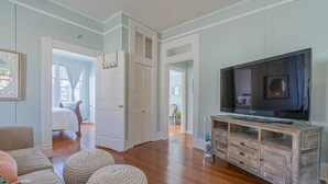 A spacious living room with a large television, wooden cabinets, and a cozy seating area with ottomans. - Walk to Beach & UTMB |Dogs Ok |Game Room (Galveston)