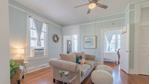 A living room with a beige sofa, a clear coffee table, and a round pouf. There is a ceiling fan, a window with curtains, and a door leading to another room. - Walk to Beach & UTMB |Dogs Ok |Game Room (Galveston)