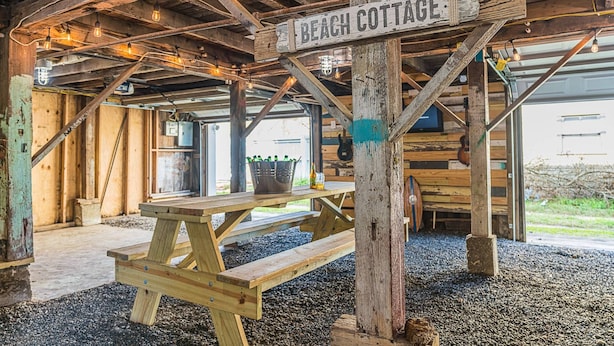 A rustic wooden structure labeled "Beach Cottage" with exposed beams, a wooden bench, and a table with bottles. - Walk to Beach & UTMB |Dogs Ok |Game Room (Galveston)