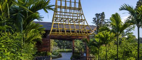 Una estructura de pérgola de madera con techo inclinado, rodeada de abundante vegetación y palmeras.