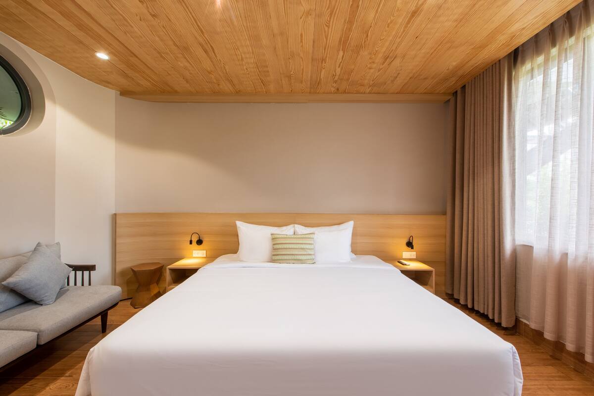 Une chambre d’hôtel avec un grand lit, un plafond en bois, deux tables de chevet avec des lampes, une fenêtre ronde et un canapé.