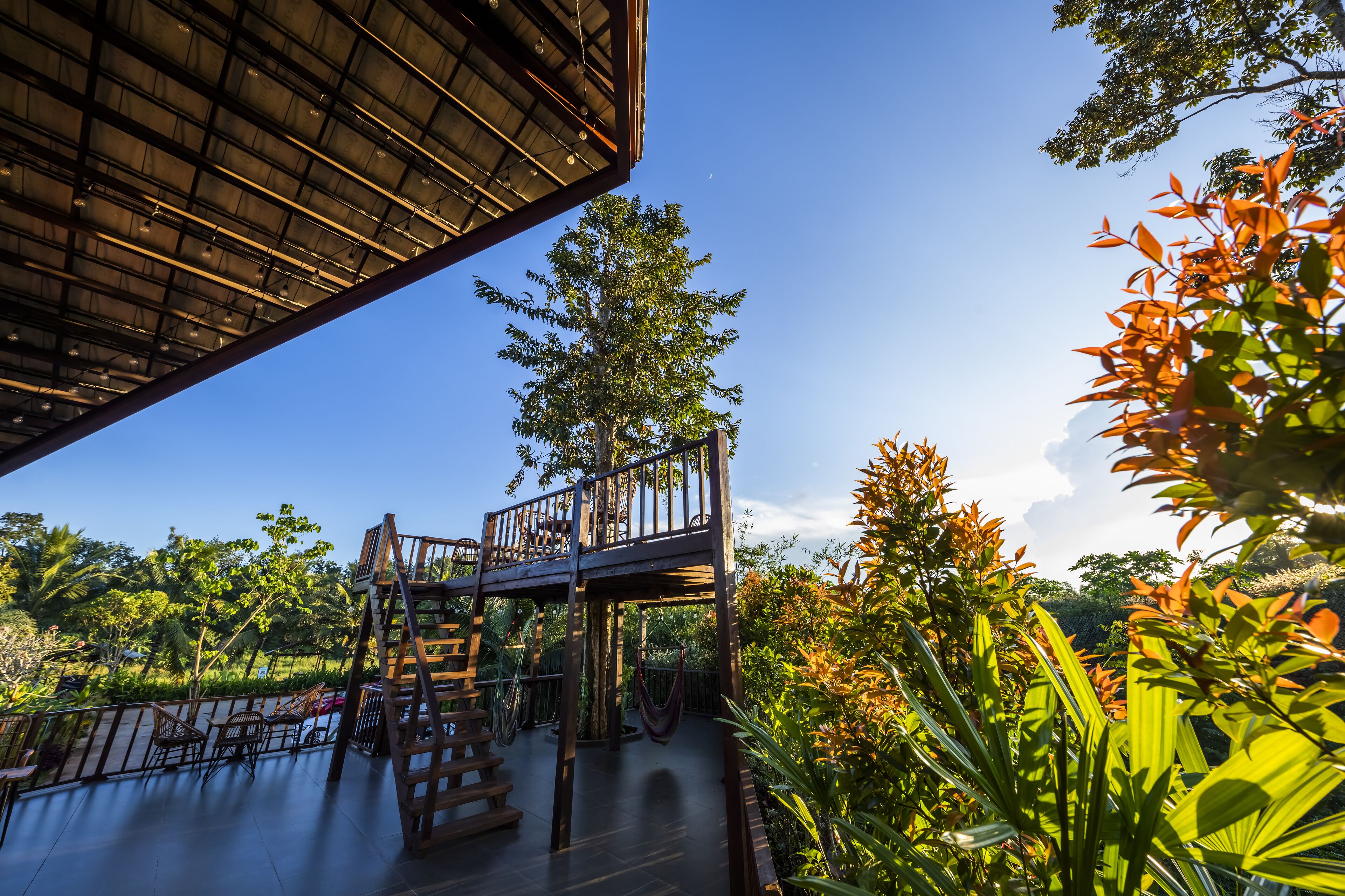 A wooden deck with chairs and a hammock, surrounded by lush greenery and a clear blue sky.