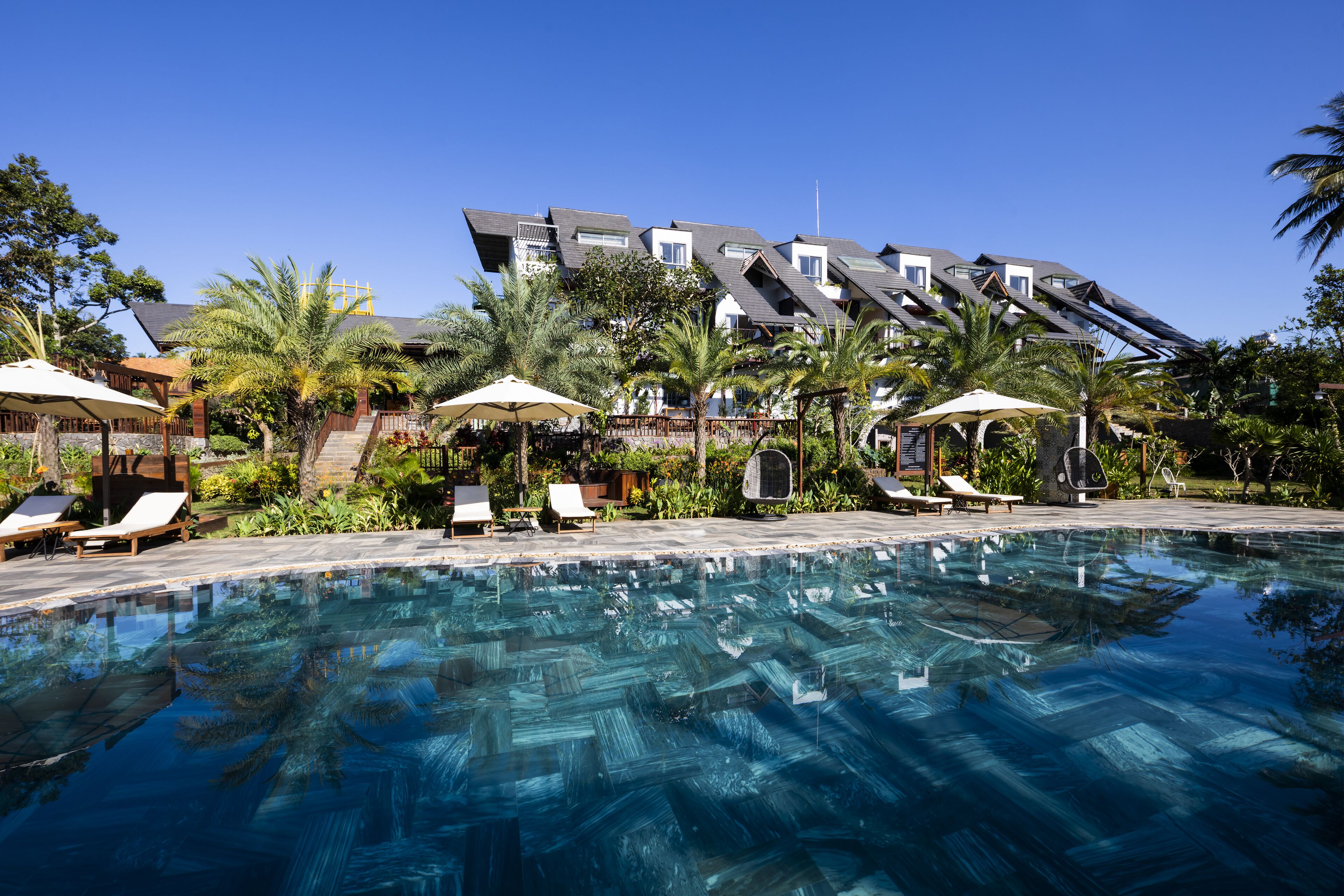 A swimming pool with lounge chairs and umbrellas, surrounded by palm trees and a building with a sloped roof.