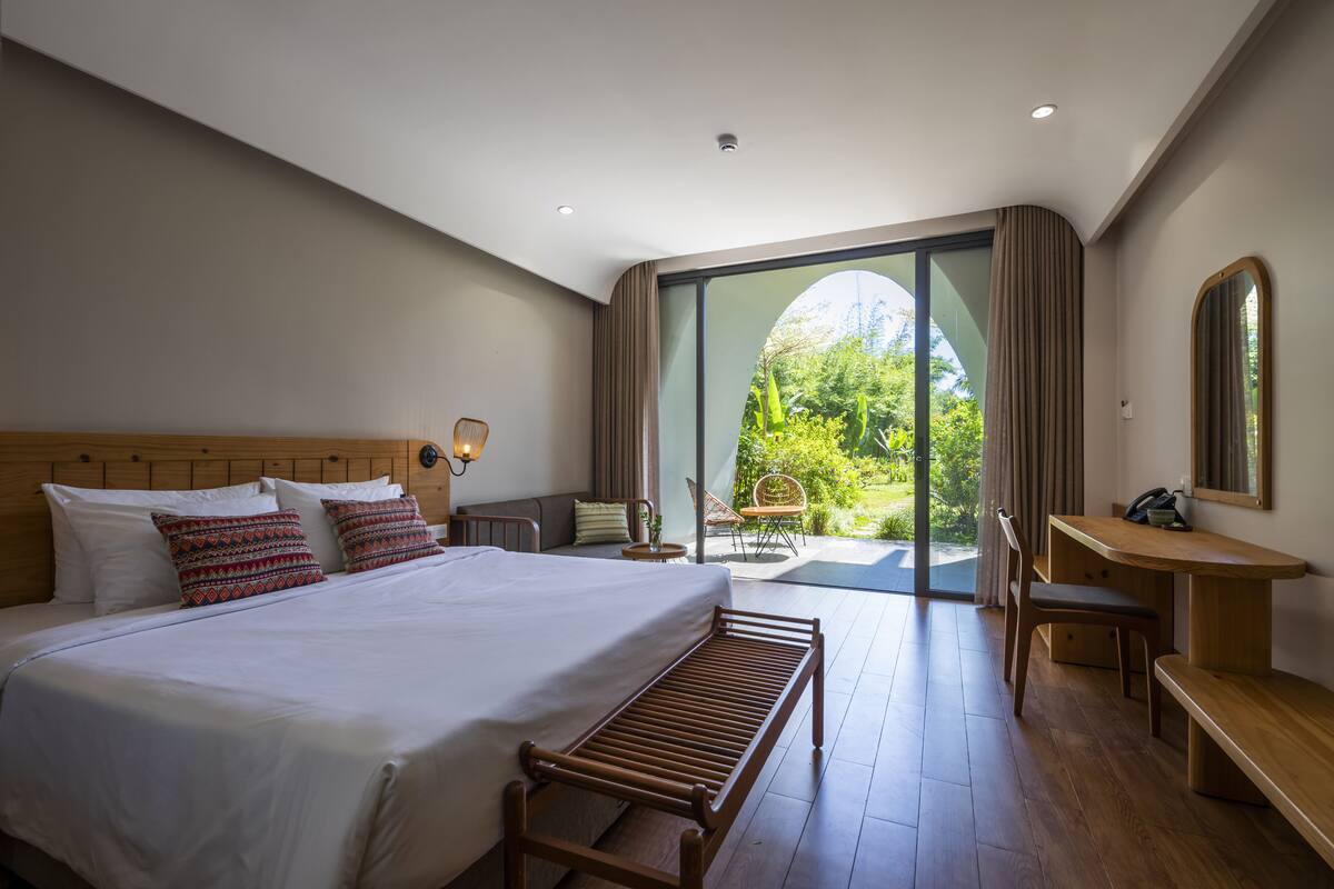 Une chambre d’hôtel moderne avec un grand lit, un banc en bois, un bureau et une chaise. Une porte-fenêtre coulissante donne accès à un espace extérieur verdoyant.