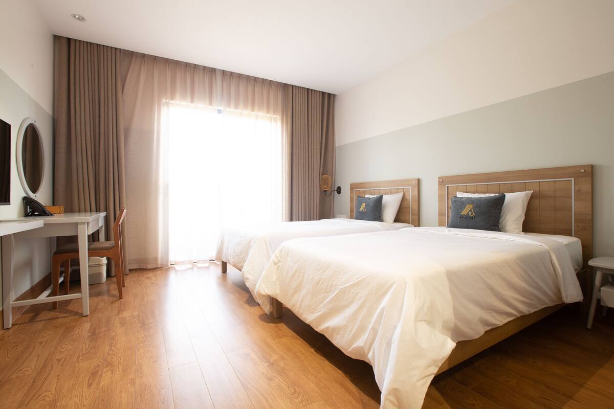 Une chambre d’hôtel avec deux lits, un bureau avec une chaise, une petite table et une grande fenêtre avec des rideaux.