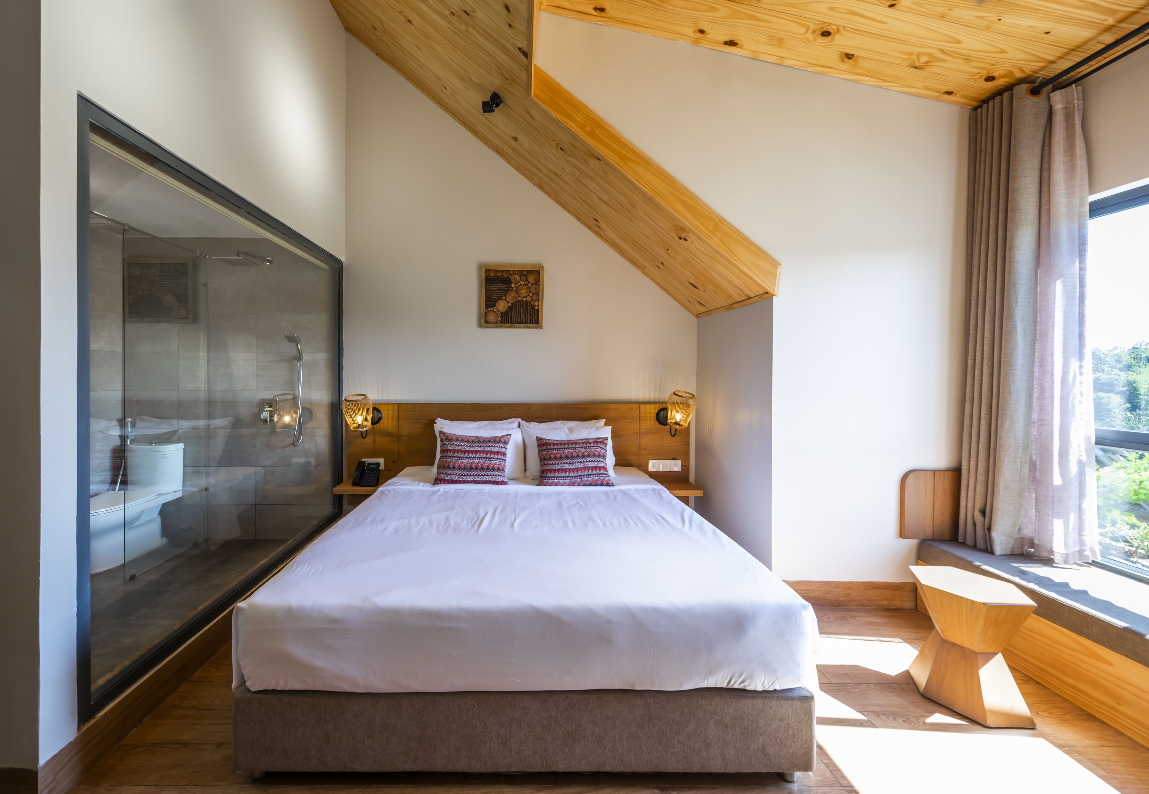 A modern bedroom with a large bed, a glass shower enclosure, wooden beams on the ceiling, and a view of greenery outside.