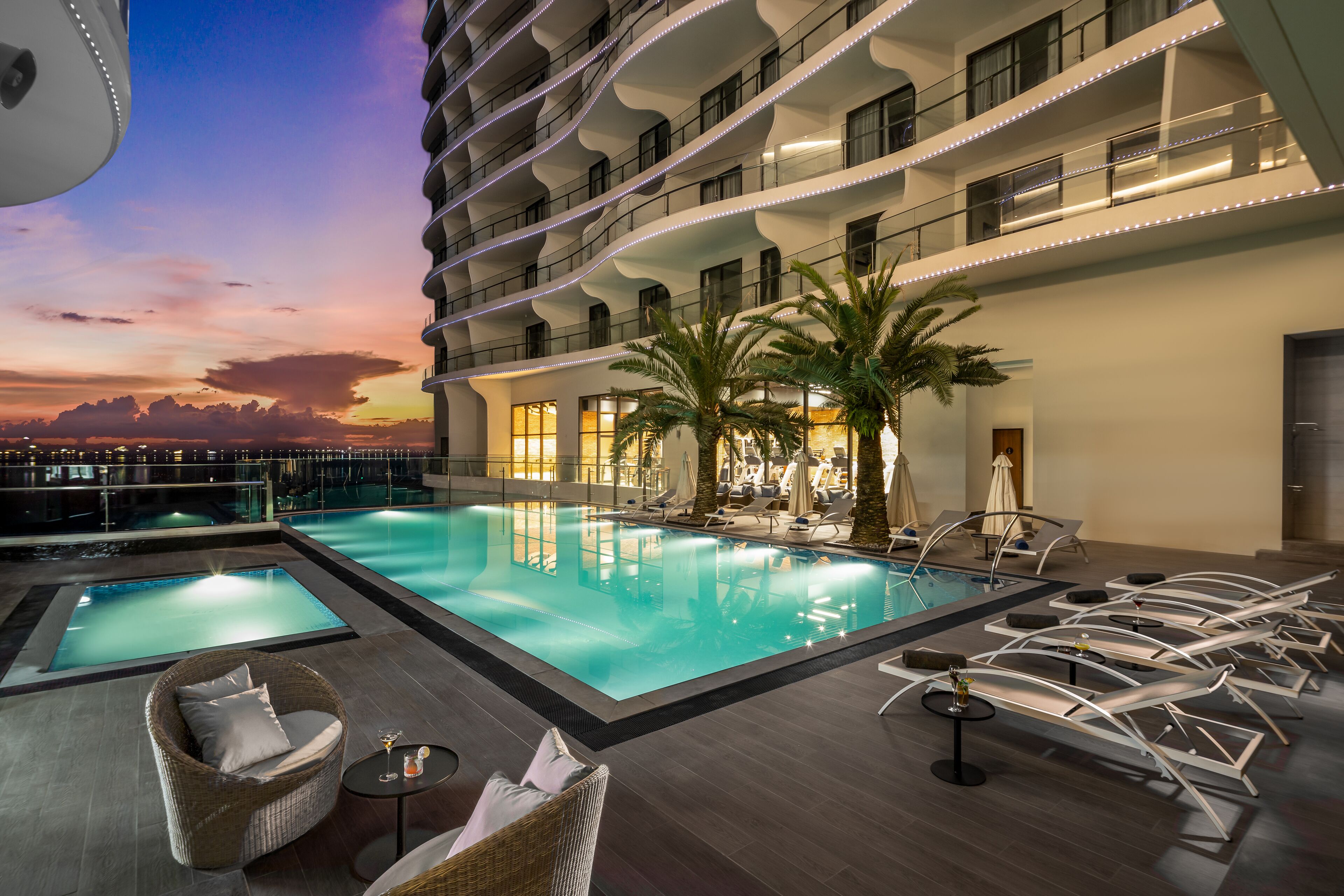 A rooftop pool area with lounge chairs, a hot tub, and a view of the sunset.