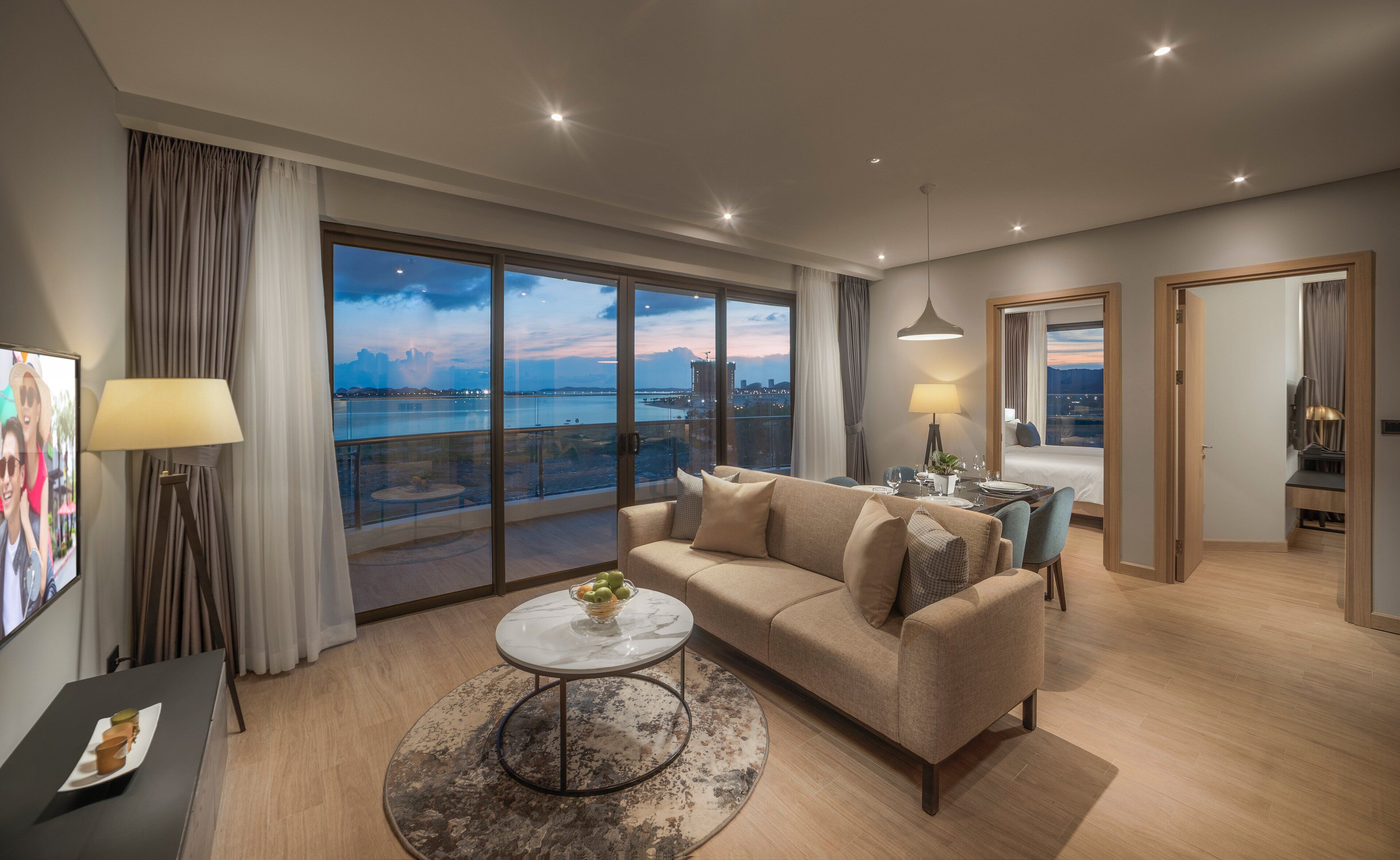A modern hotel room with a large balcony, a sofa, a dining table, and a TV.