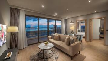 A modern hotel room with a large balcony, a sofa, a dining table, and a TV.