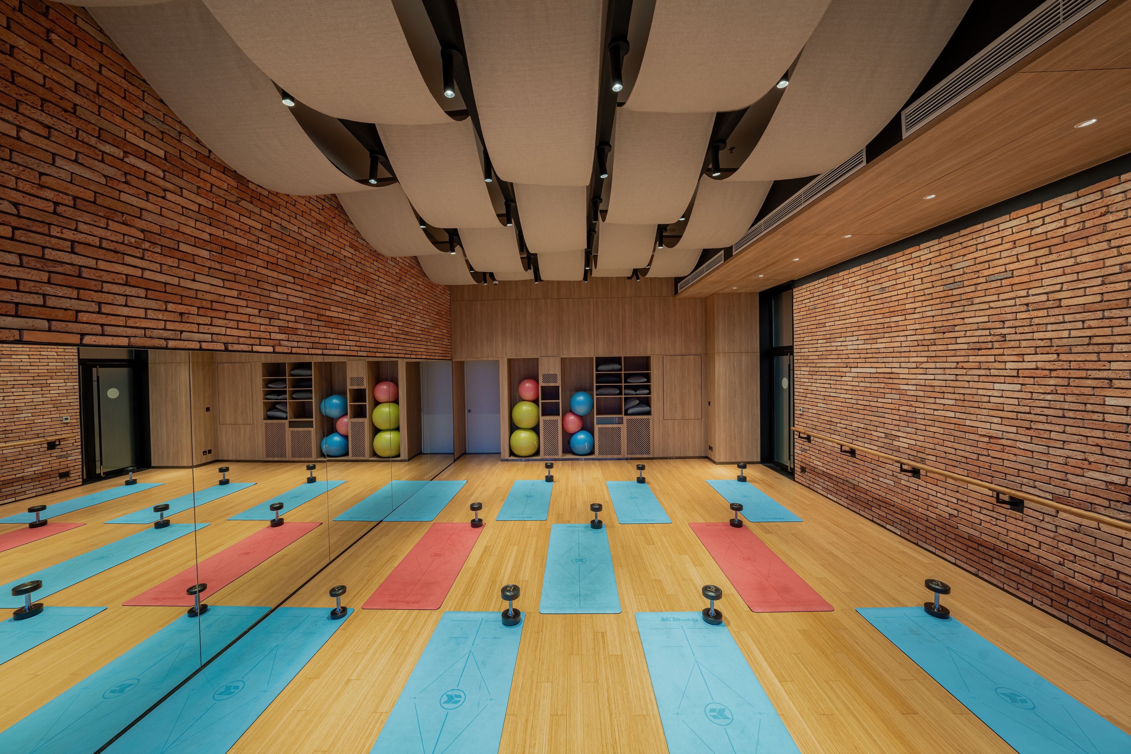 Une salle de sport spacieuse, dotée de nombreux tapis et appareils d’exercice, de planchers en bois, de murs en briques et d’un plafond haut avec des spots encastrés.