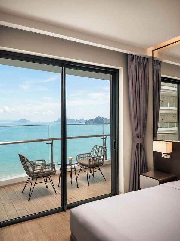 A hotel room with a balcony, a bed, a chair, a table, and a view of the sea. - Citadines Marina Halong (Bai Chay)