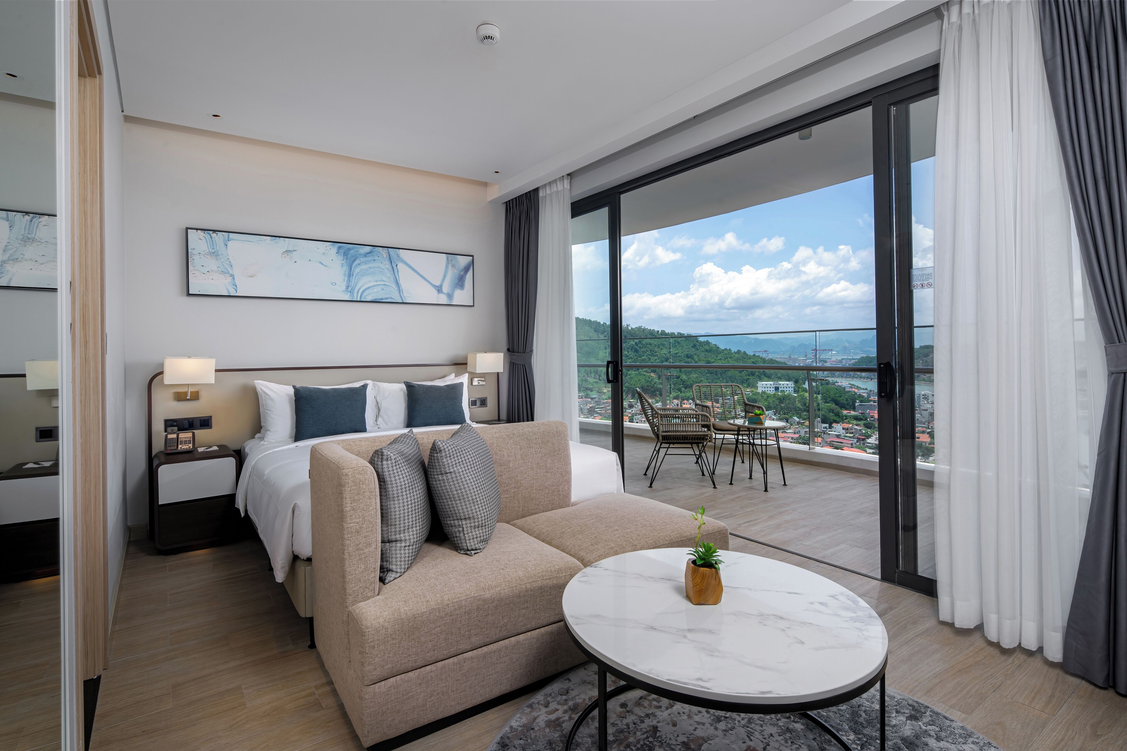 A modern hotel room with a bed, a sofa, a small table with a plant, and a balcony with a view of the city.