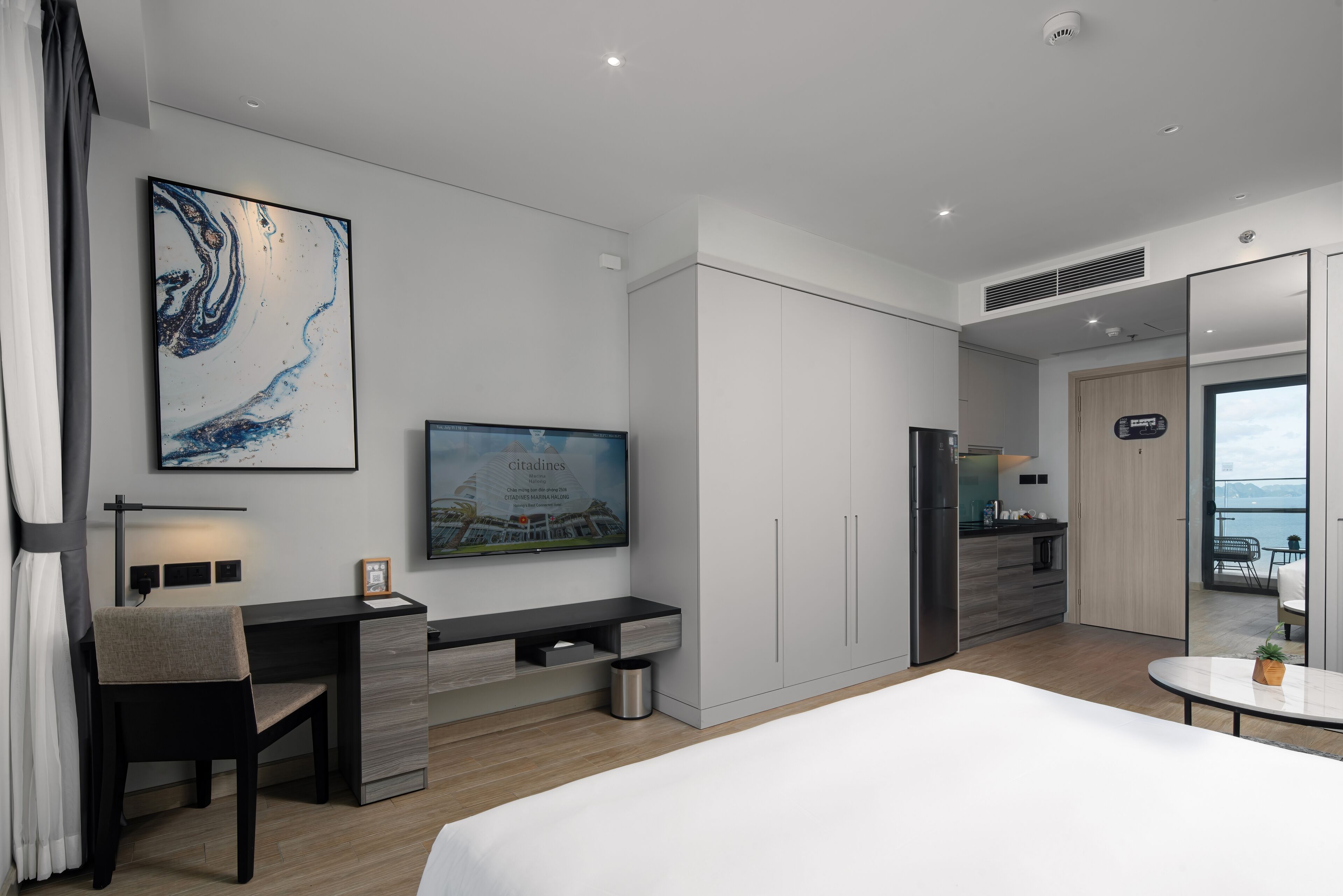 Moderni hotellihuone, jossa on työpöytä, tuoli, sänky ja suuri ikkuna, josta avautuu näkymä merelle.
