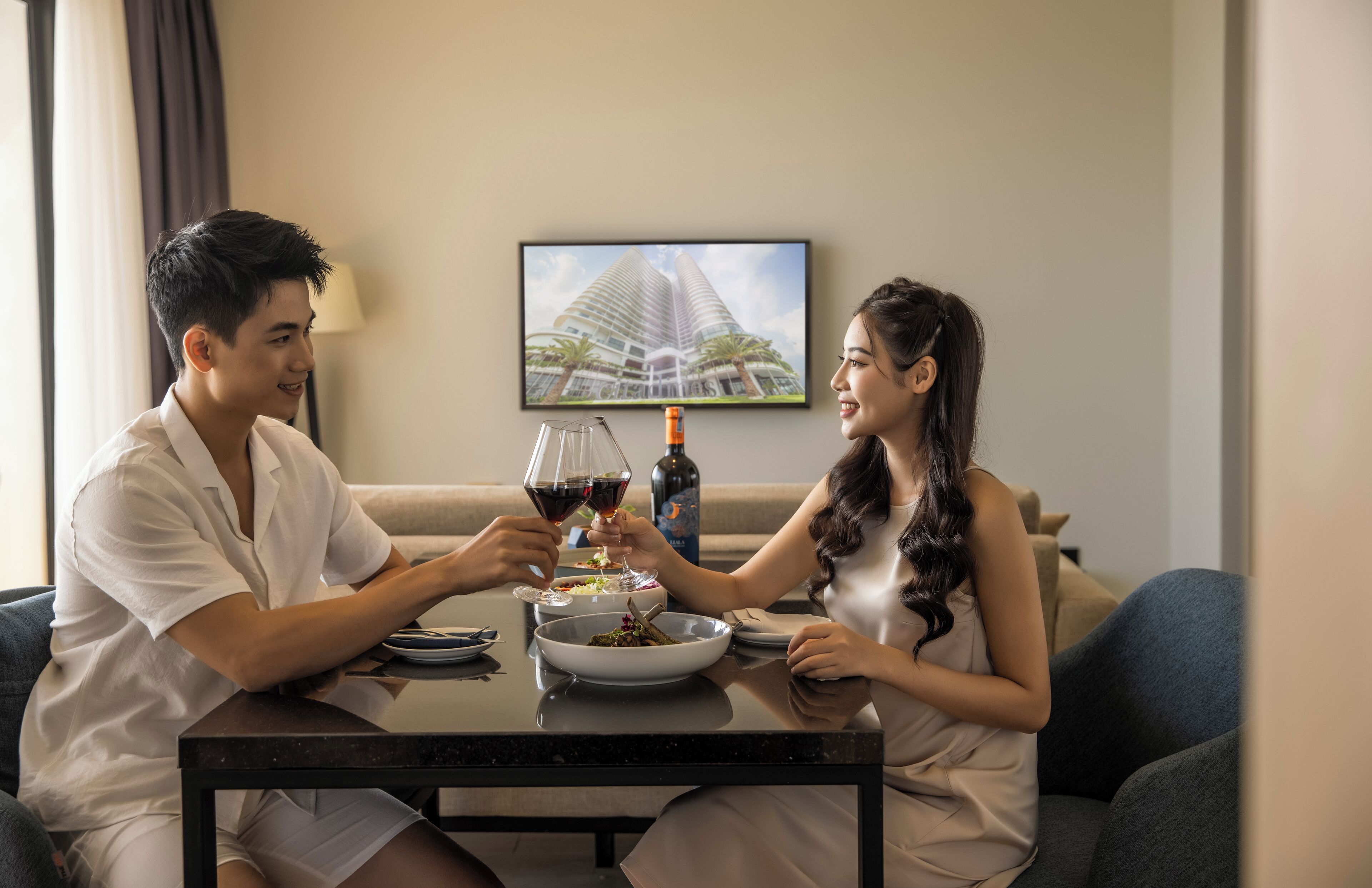 A couple seated at a table with a bottle of wine, a bowl of food, and a framed picture of a building on the wall.