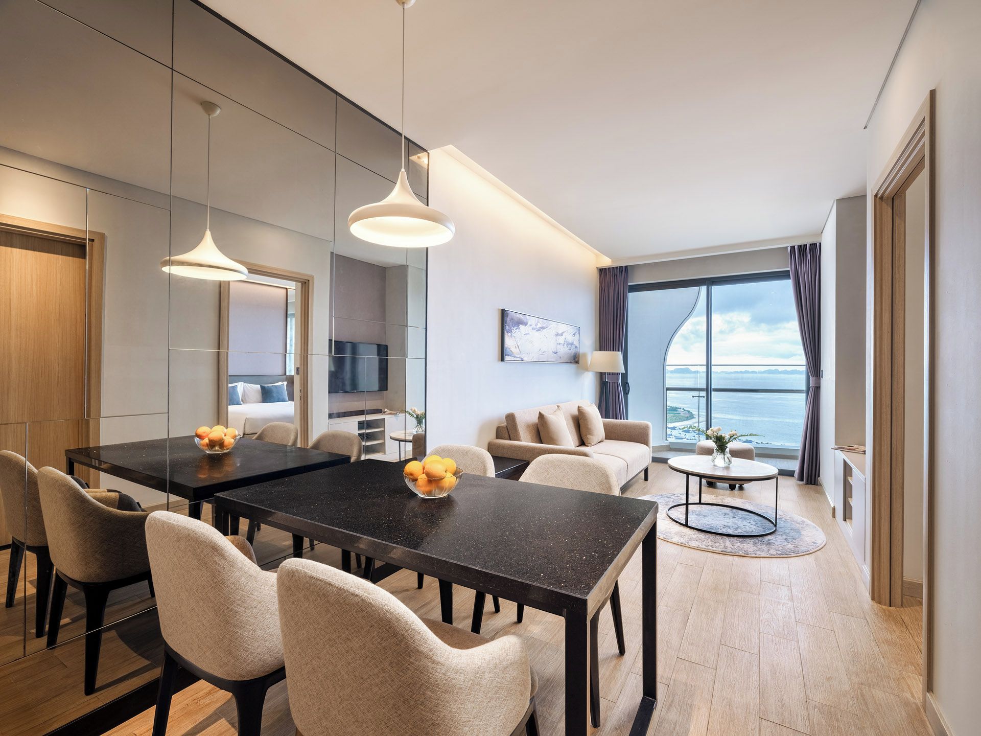 A modern hotel room with a dining area, a sofa, a TV, and a view of the sea.