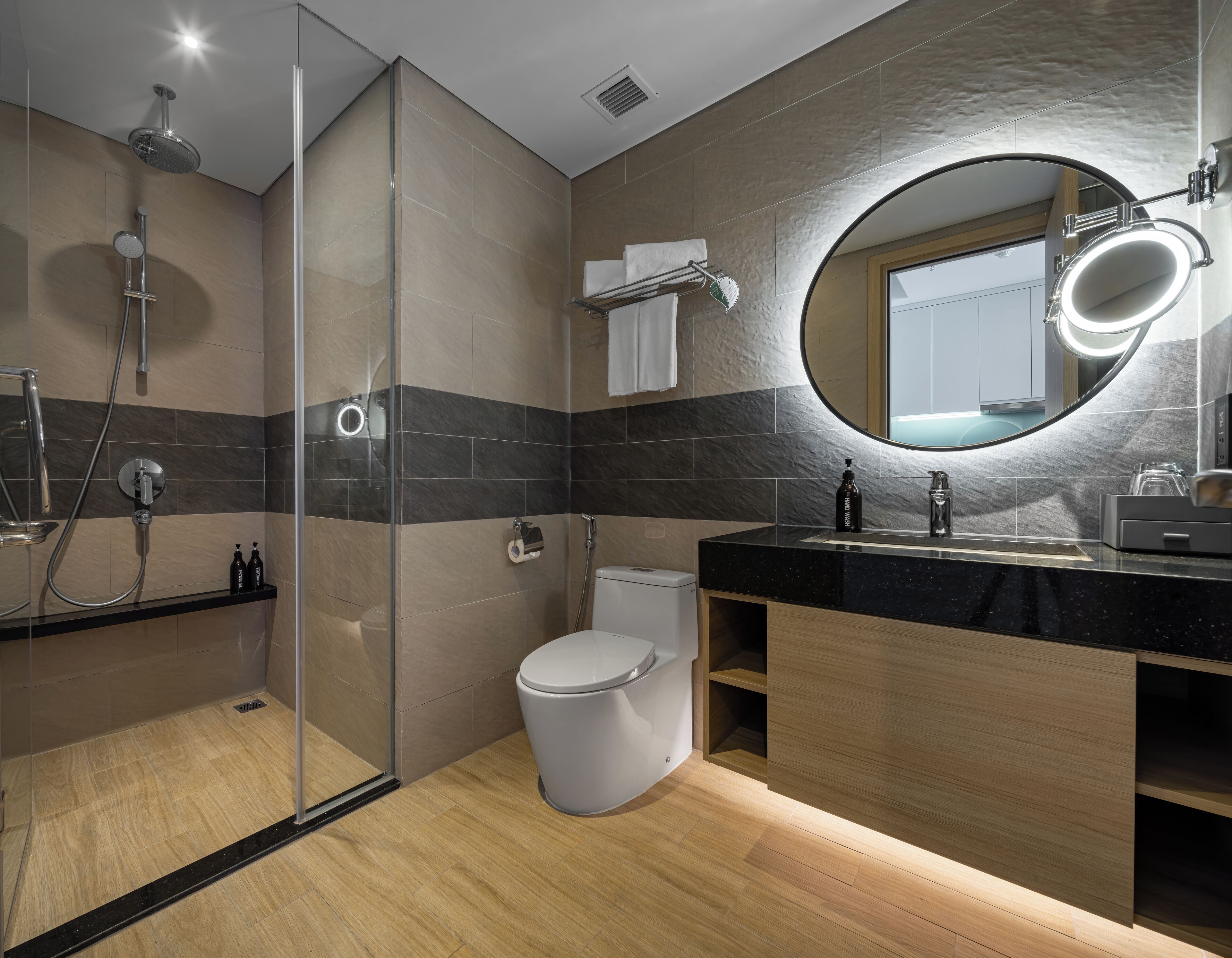 Egy modern fürőszoba üveg válaszfallal ellátott zuhanyzóval, WC-vel, tükörrel ellátott mosdókagylóval, valamint törölközőtartóval rendelkező mosdóasztallal.