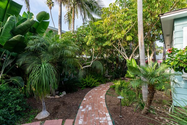Un sendero de ladrillos que atraviesa un jardín exuberante con diversas plantas tropicales y palmeras.