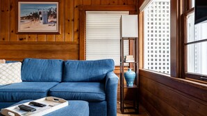 Una habitación con un sofá azul, una bandeja con mandos a distancia, un cuadro enmarcado y una ventana con persianas.