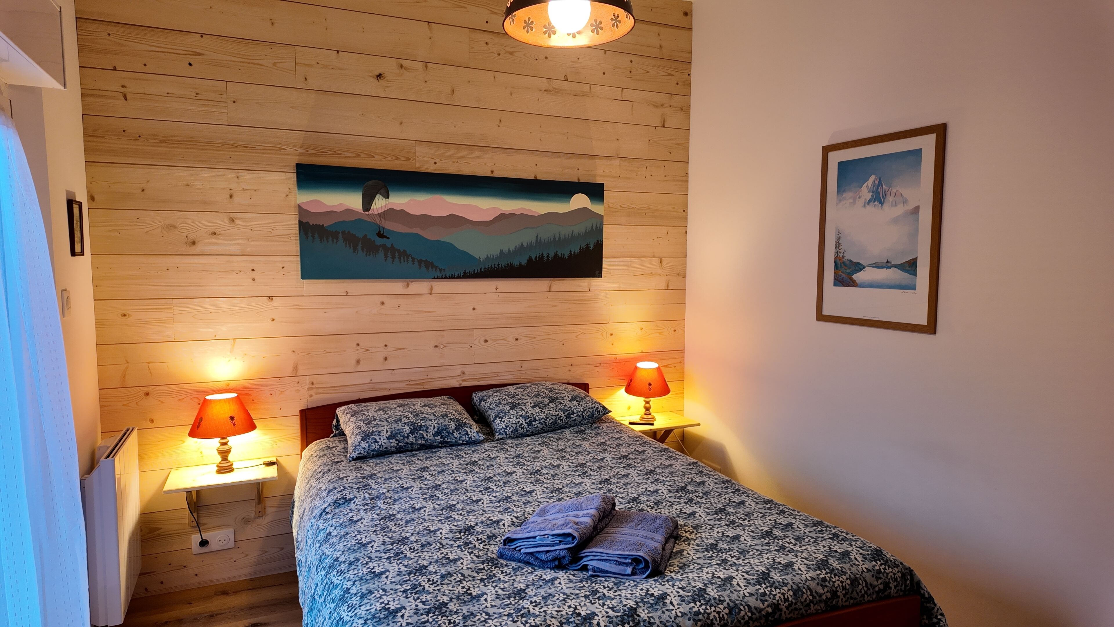 Une chambre avec un plafond en bois, un lit recouvert d’une couvre-lit à motifs, deux lampes de chevet et deux tableaux encadrés accrochés au mur.