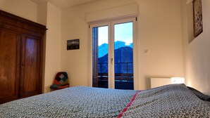 A bedroom with a bed, a wooden wardrobe, a chair, a window with a mountain view, and a wall-mounted light fixture. - New Spacious Appart. Incredible view of Mt Blanc (Passy)