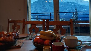 A dining table with a fruit bowl, a loaf of bread, and a mug, with a mountain view through large windows. - New Spacious Appart. Incredible view of Mt Blanc (Passy)