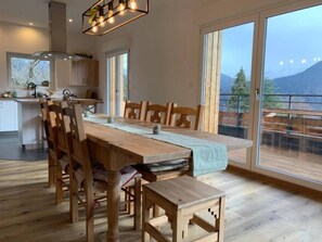 A spacious dining area with a wooden table and chairs, large windows offering a mountain view, and a modern kitchen in the background.