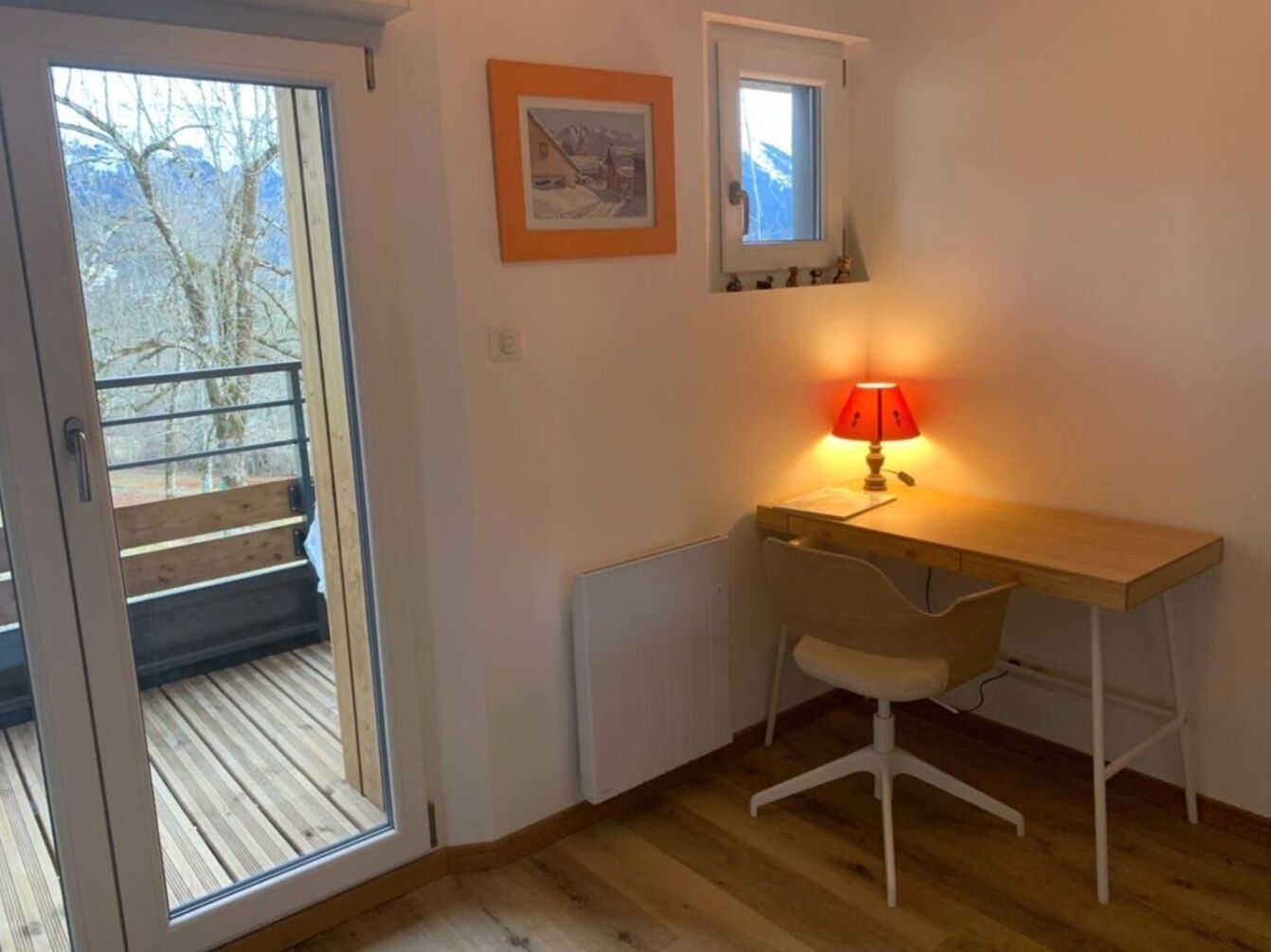 A room with a wooden desk, a chair, a red lamp, a radiator, a window, and a door leading to a balcony.