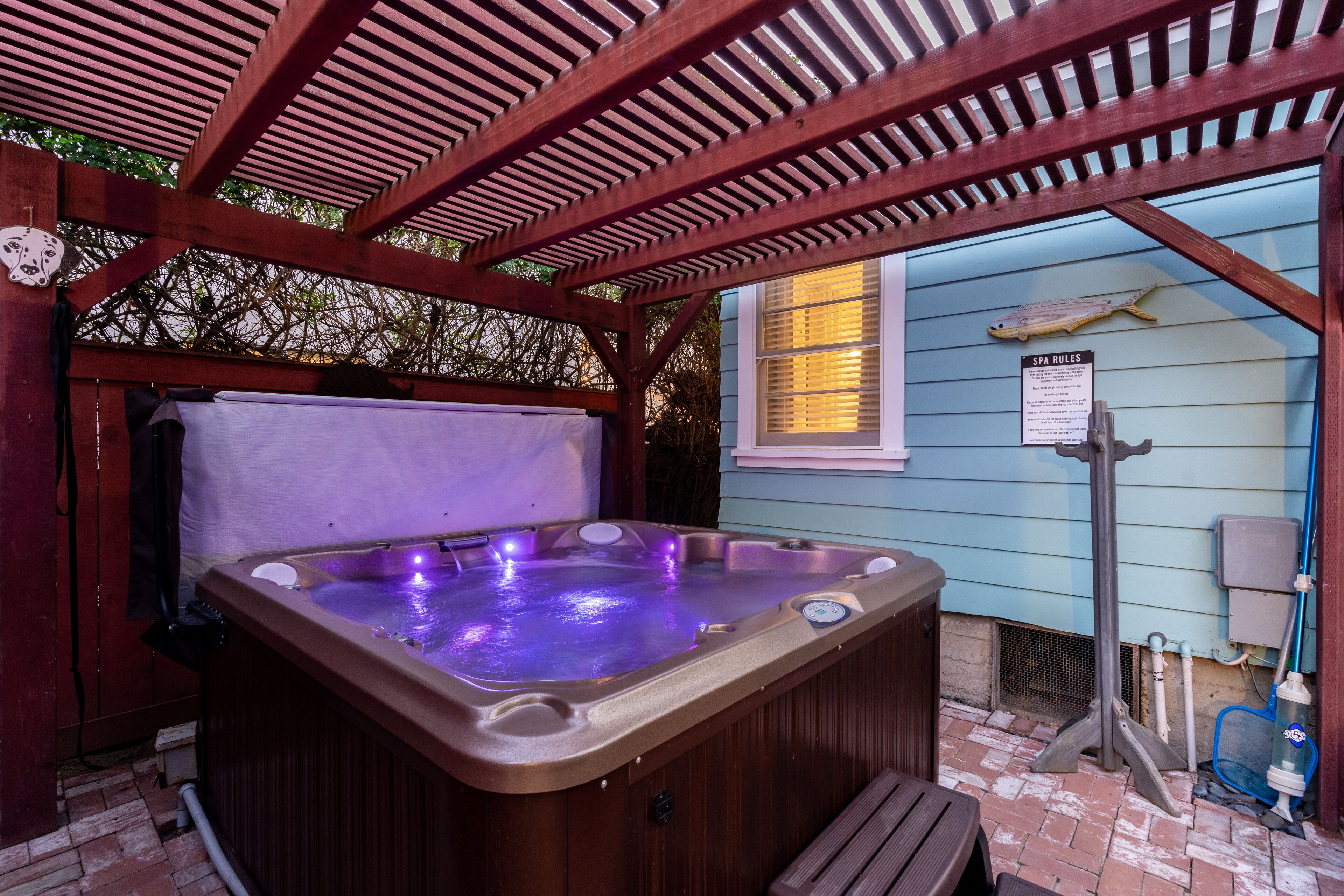 Un bain à remous sous une pergola en bois, avec un panneau indiquant les règles d'utilisation du spa, une décoration en forme de poisson et une maison bleue en arrière-plan.