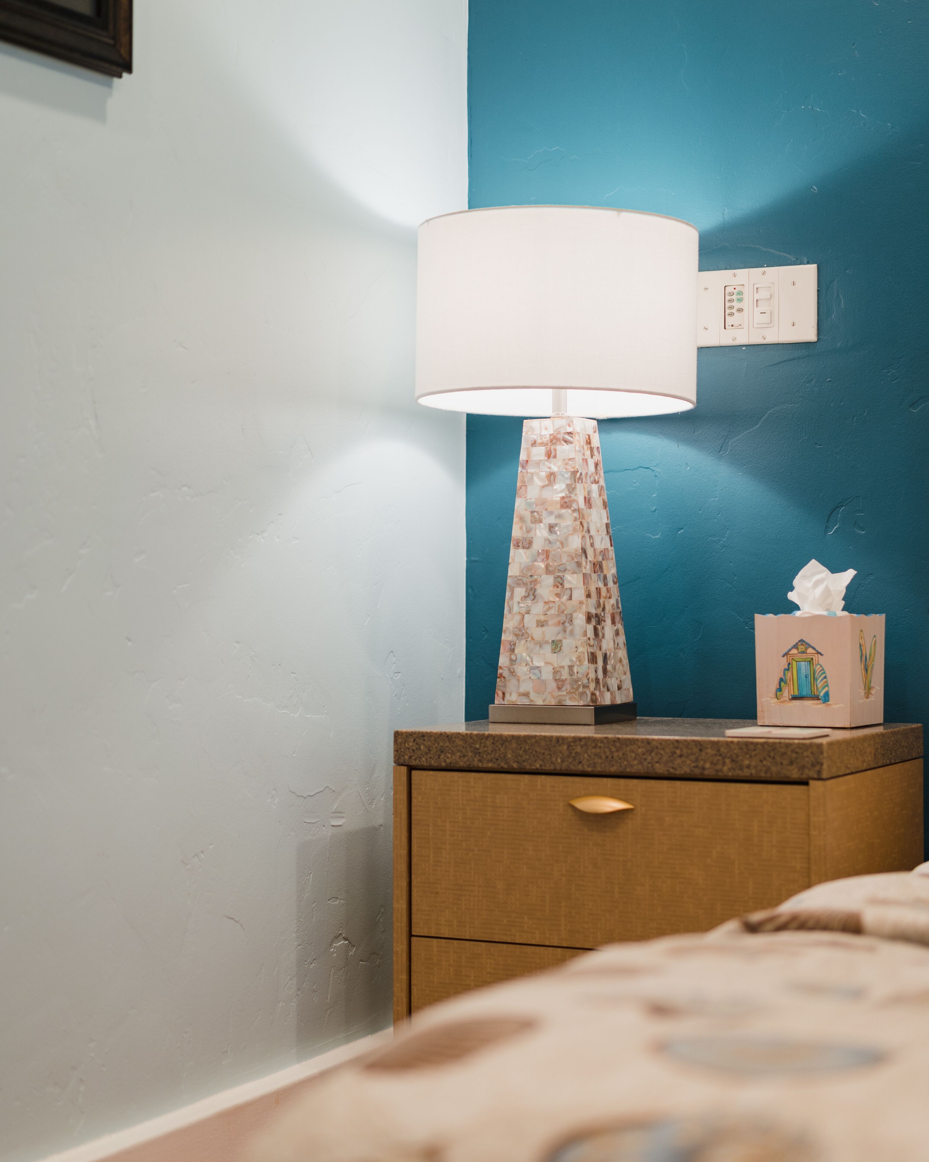 A bedside table with a lamp, a tissue box, and a power outlet on the wall.