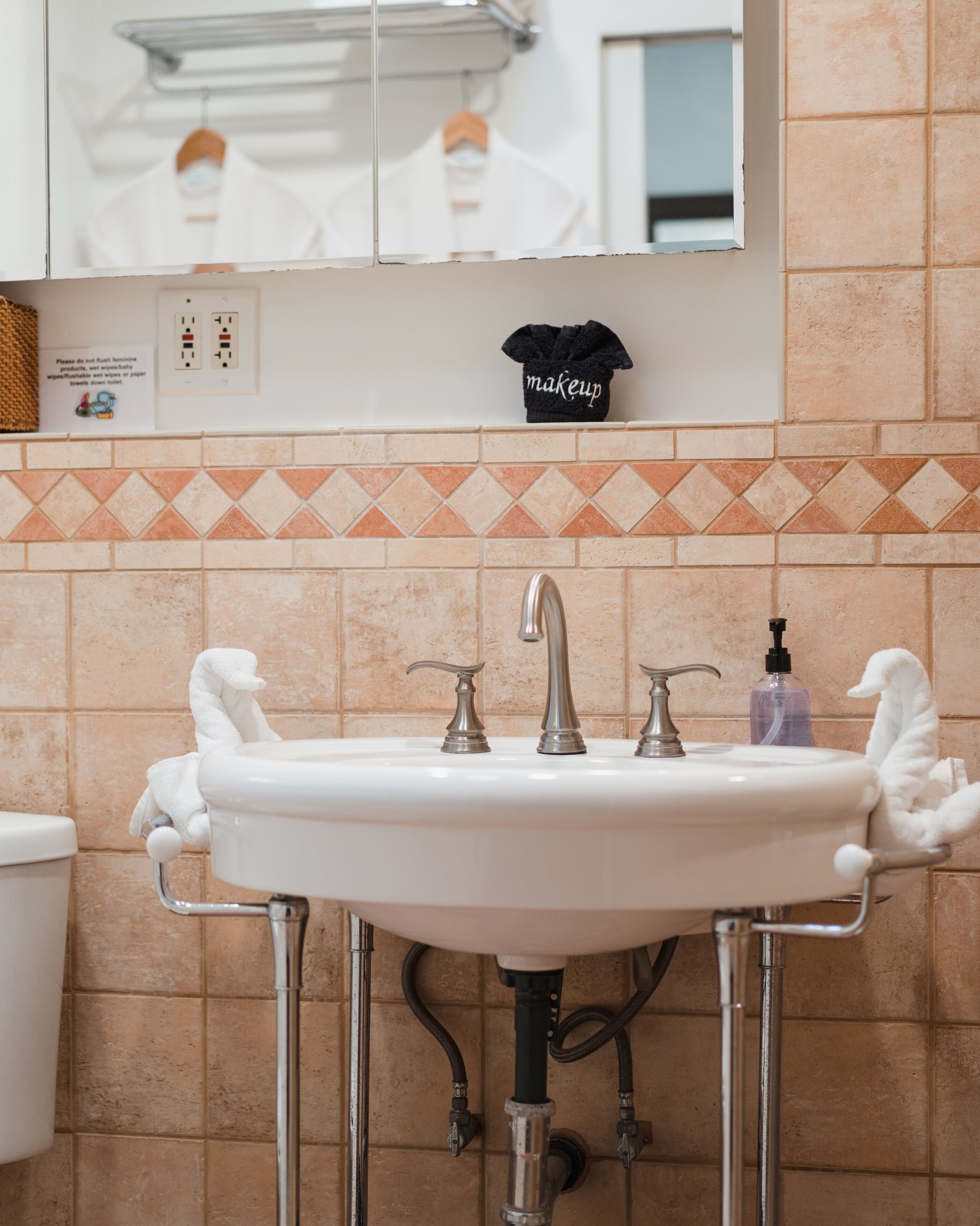 Une salle de bain avec un lavabo blanc, deux robinets et un miroir au-dessus. On y trouve un porte-serviettes avec une serviette noire et une pancarte au-dessus.