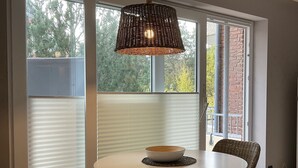 Zona pranzo con tavolo rotondo, due sedie in vimini, lampada a sospensione intrecciata e vista sugli alberi attraverso ampie finestre.