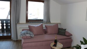 A pink sofa with a bottle of champagne and a glass on a small round table, a window with curtains, and a radiator below. - Relaxation in the Ammergau mountains, centrally located (Oberammergau)