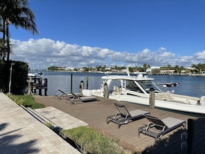 Relax on the waterfront with a docked boat and comfortable sun loungers on the deck, surrounded by lush greenery and palm trees.