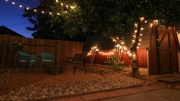 Een achtertuin met een terras, lichtjes, een houten schutting en een tuinhuis.