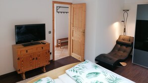 Un dormitorio con una cama, un televisor sobre un mueble de madera, una silla y una puerta que da a otra habitación.